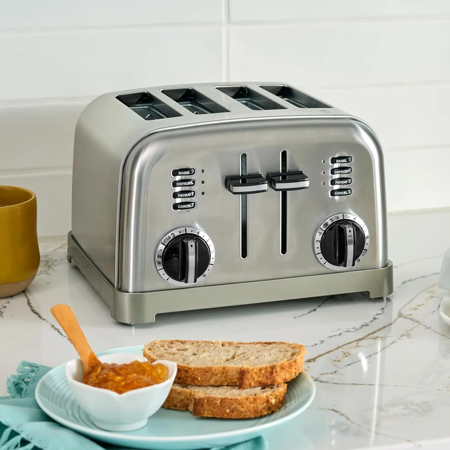 Brushed stainless steel Cuisinart four-slice toaster on a marble counter with bread and a bowl of jam